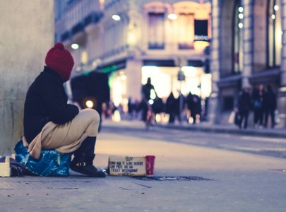 A homeless person sitting on a street corner as people walk by